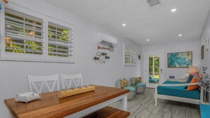 Una habitación con una mesa de comedor de madera, sillas blancas, una cama con ropa de cama color turquesa y una ventana con persianas.