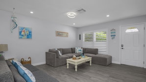 Una sala de estar con un sofá gris, una mesa de centro blanca y un sillón gris. Hay un cuadro en la pared y una puerta con un ventanal.