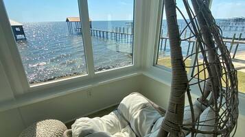 A room with a hanging chair by a large window overlooking a pier and the sea.