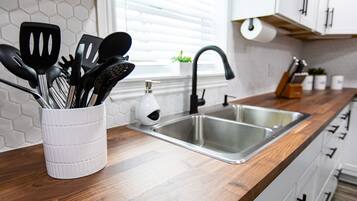 A kitchen with a stainless steel sink, a white utensil holder, and a window with blinds.