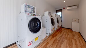 A laundry facility with multiple washing machines lined up against a wall, a wooden floor, and a vent on the ceiling. - HOTEL R9 The Yard Hitachinaka (Hitachinaka)
