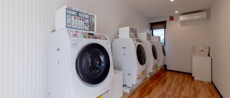 A laundry facility with multiple washing machines lined up against a wall, a wooden floor, and a vent on the ceiling.