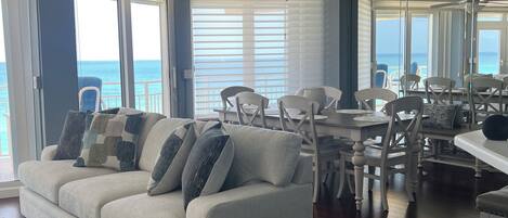 A spacious living room with a white sofa, dining area, and large windows offering a view of the ocean.