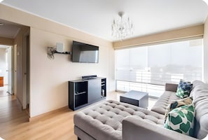 A modern living room with a flat-screen TV mounted on the wall, a sofa, a coffee table, and a large window offering a view of the city. - Barranco Duplex Penthouse (Barranco)