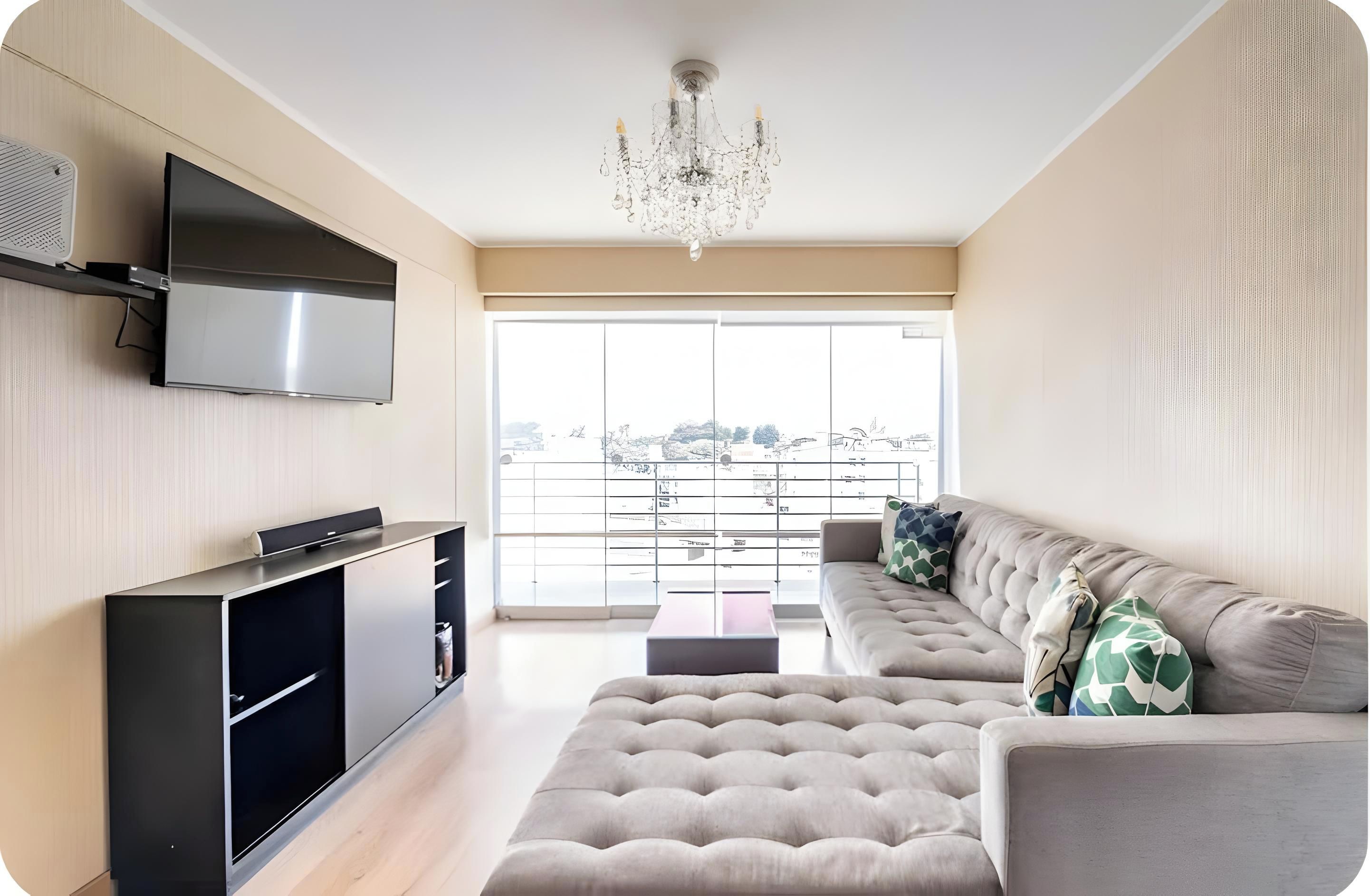 A modern living room with a flat-screen TV, a large sofa, a coffee table, and a chandelier.