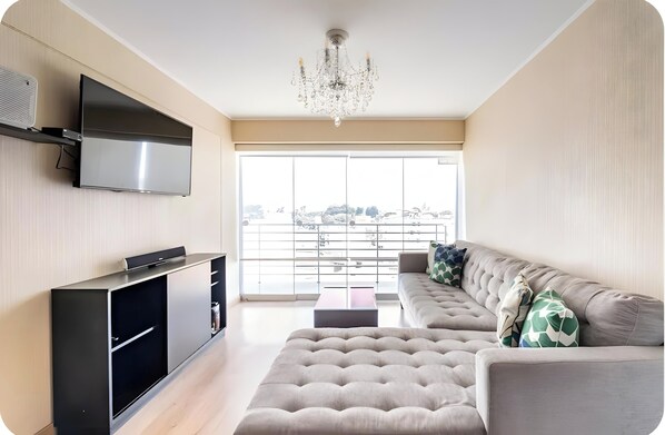 A modern living room with a flat-screen TV, a large sofa, a coffee table, and a chandelier.