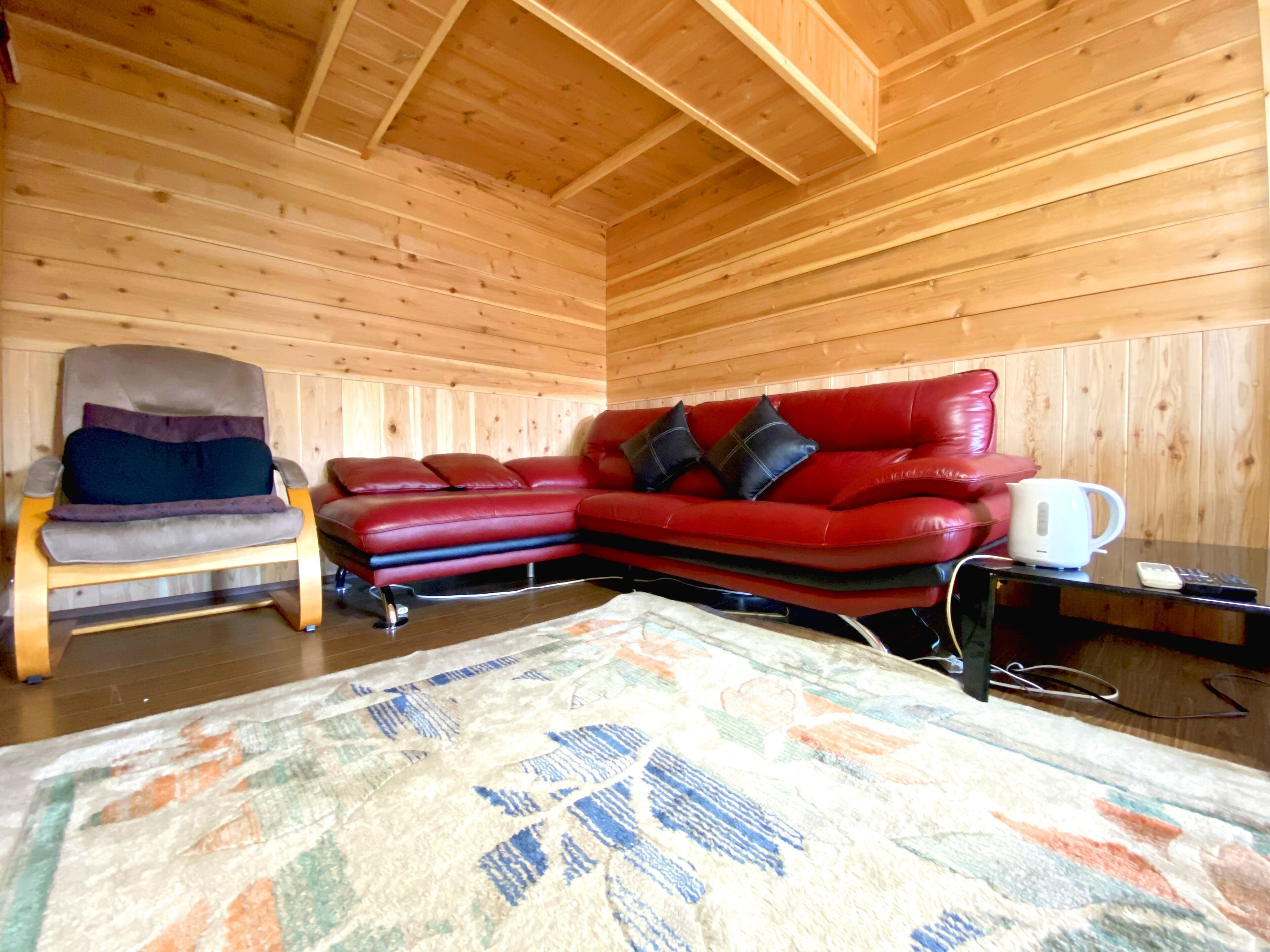 A wooden interior with a red leather sofa, a wooden chair, a small table with a kettle, and a patterned rug.