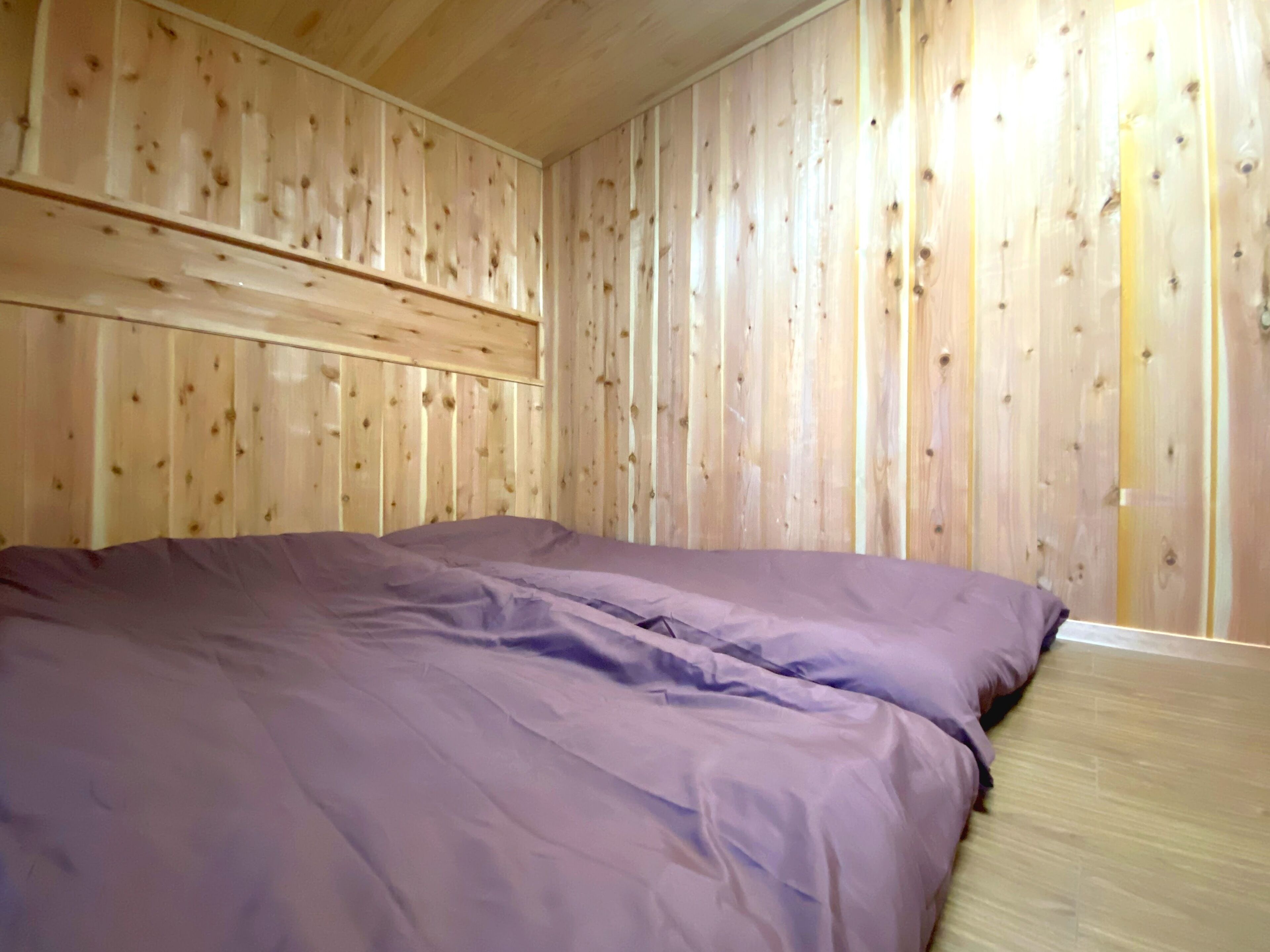 A room with wooden panel walls, a bed with purple bedding, and a wooden floor.