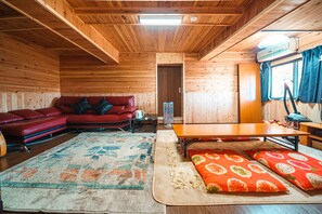 A cozy cabin interior with a wooden ceiling, a red leather sofa, a wooden dining table, and a large rug.