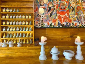 A room with wooden shelves displaying numerous Buddha statues, a large colorful mural, and white candle holders with candles on the floor.