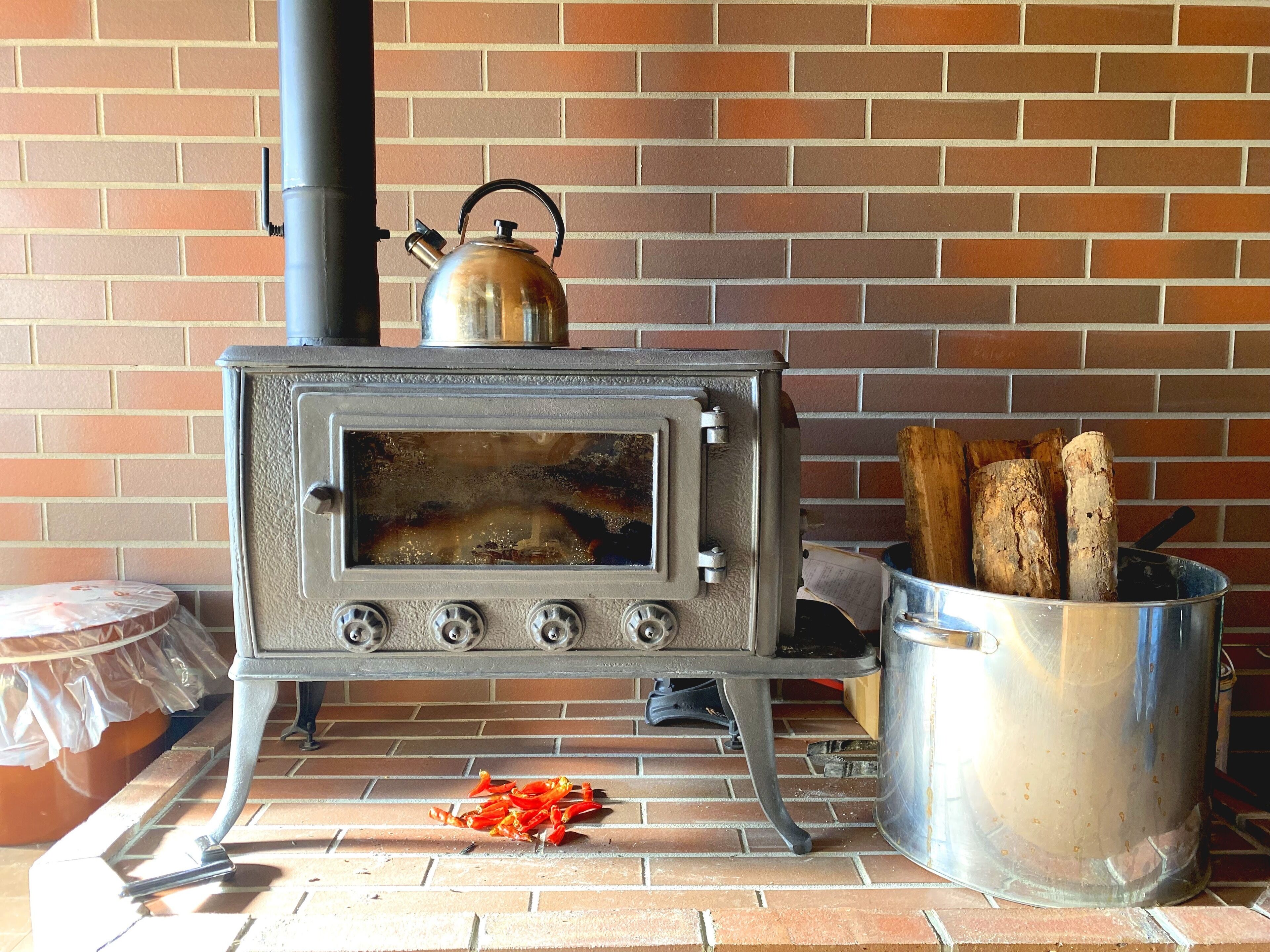 Un horno a leña con un hervidor encima, un cubo de metal con leña, y un chile rojo en el suelo.