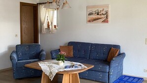 En stue med en blå sofa med et mønster, et træ sofabord med et blonde borddække, en potteplante og en blå måtte på gulvet.