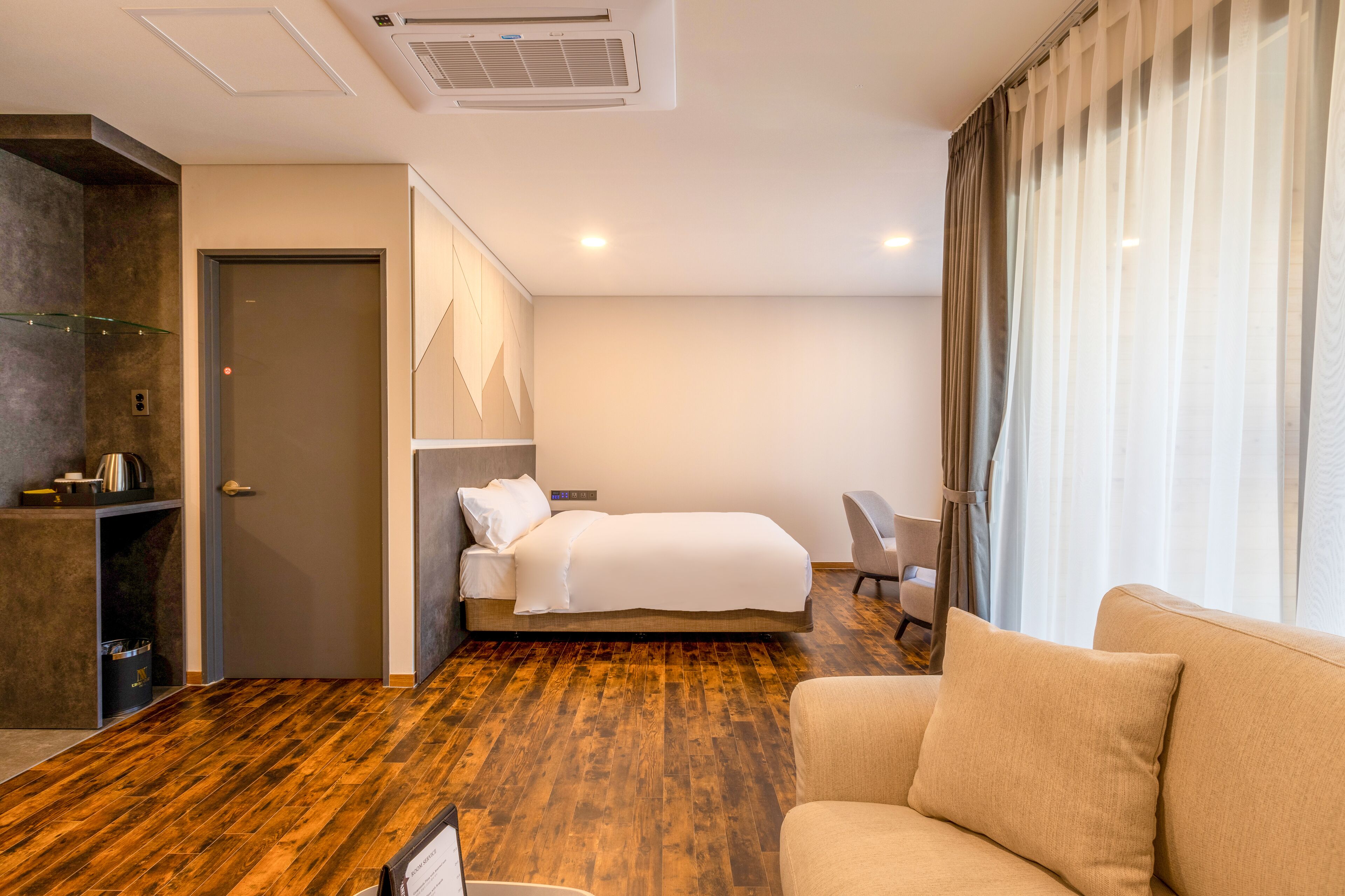 A hotel room with a wooden floor, a bed, a sofa, a desk, and a window with curtains.