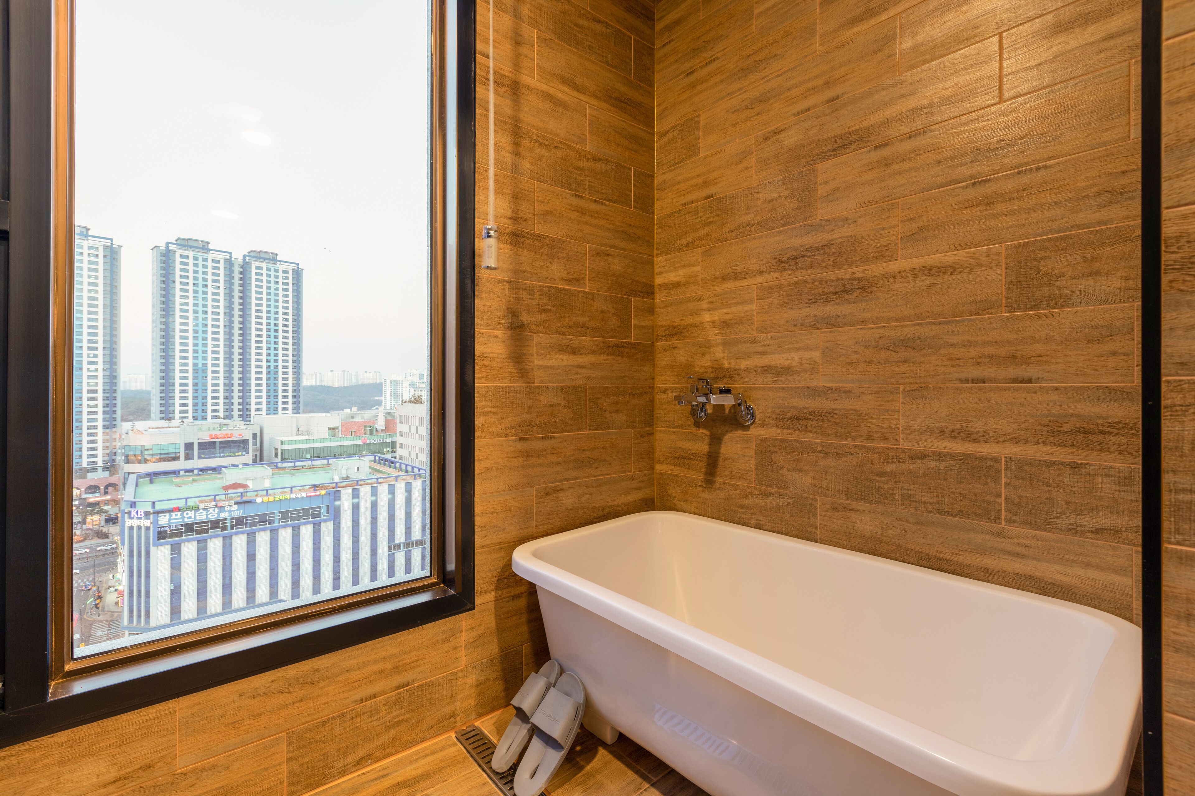 A modern bathroom with a bathtub, wooden walls, and a window offering a city view.