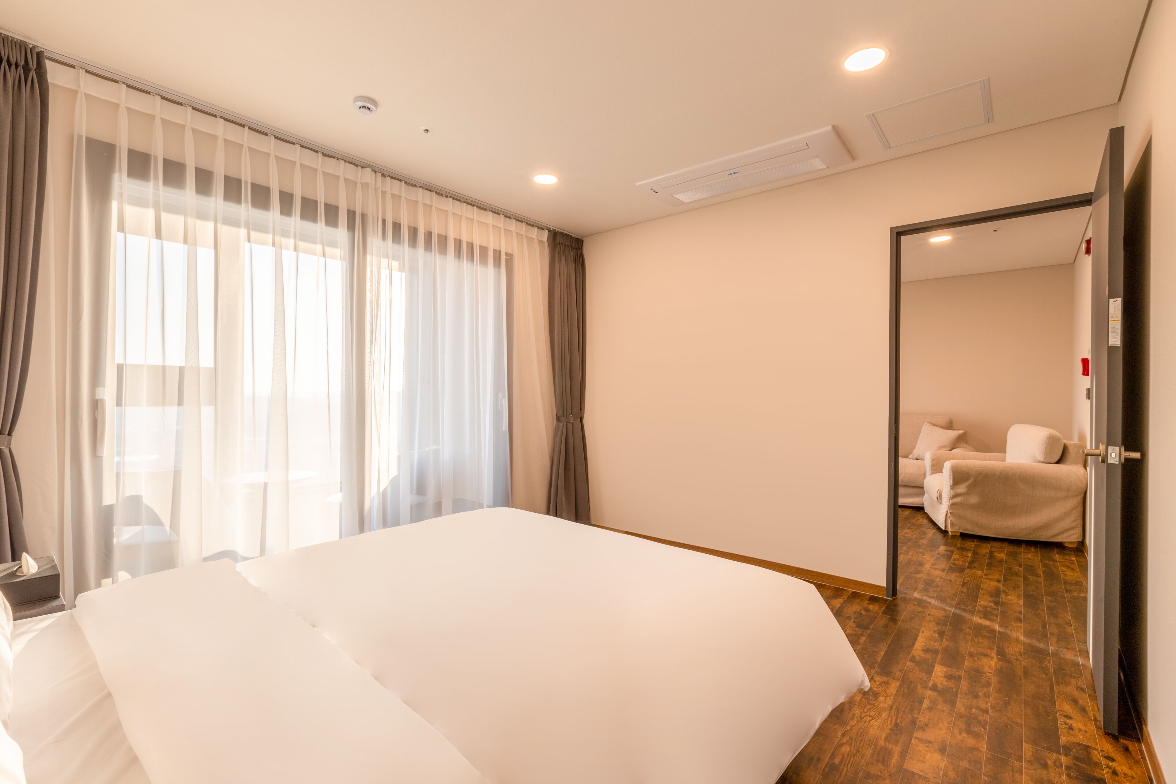 A hotel room with a large bed, a bedside table, a chair, a desk, and a view of another room.