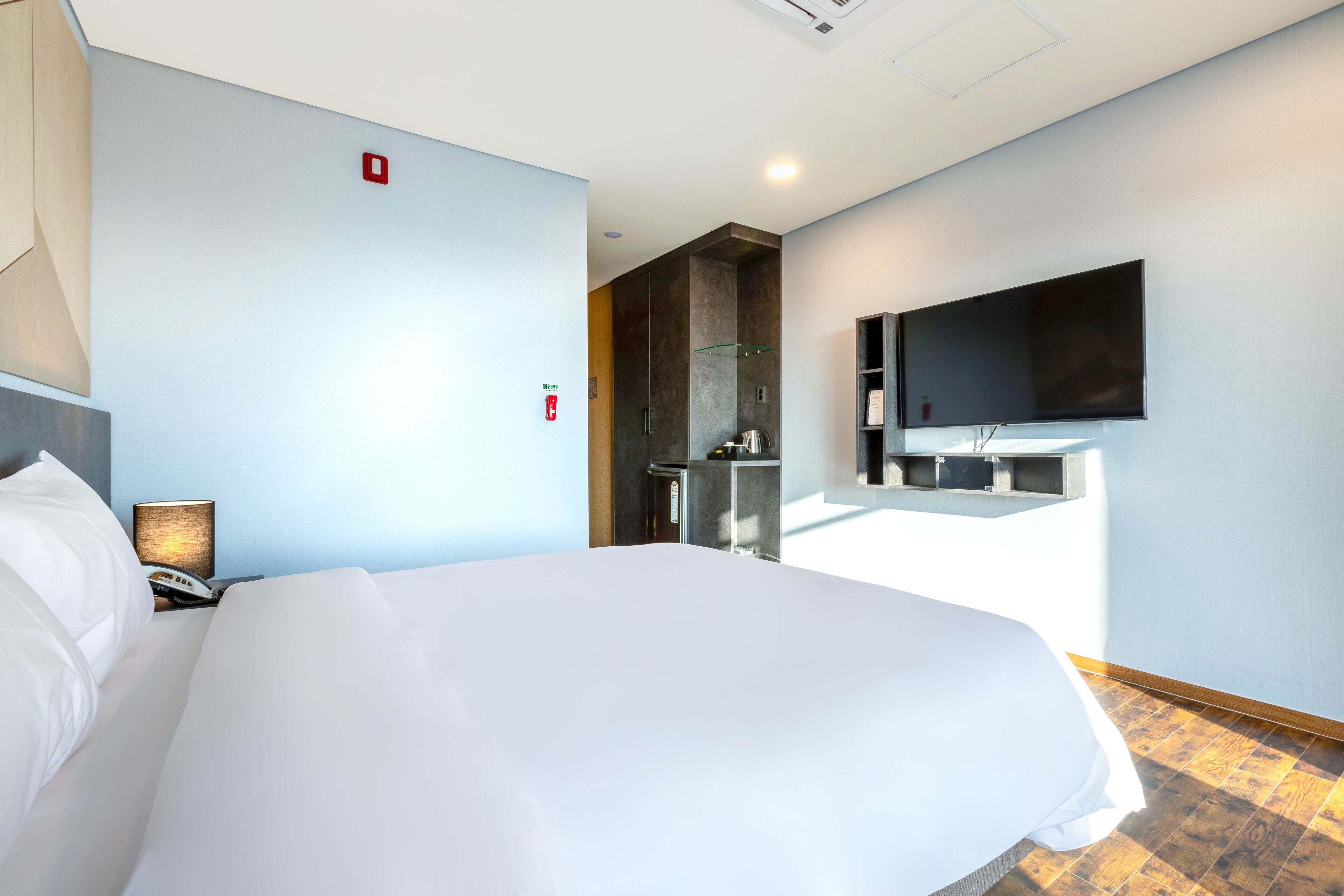A hotel room with a large bed, a flat-screen TV mounted on the wall, a bedside table with a lamp, and a shower area visible through a glass partition.