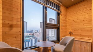 Ein modernes Zimmer mit Holzwänden und großen Fenstern, ausgestattet mit einem runden Tisch, zwei Stühlen, einem Heizkörper und Blick auf Stadtgebäude.