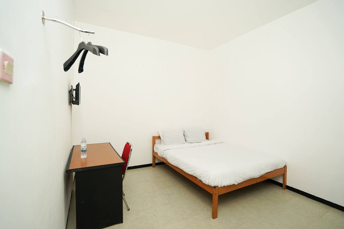 A small, clean room with a wooden bed, a desk, and a chair. There is a water bottle on the desk.