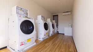 A row of four front-loading washing machines in a laundry room with wooden flooring and white walls. - HOTEL R9 The Yard Higashiomi (Higashiomi)