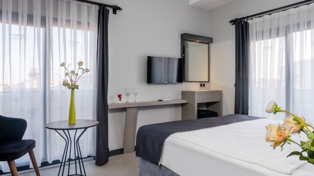 A hotel room with a bed, a small table with a vase, a TV mounted on the wall, and a desk with a mirror.