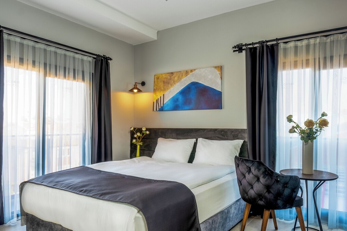 A hotel room with a bed, a chair, a small table with a vase, and a painting on the wall.