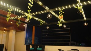 Un área de estar al aire libre con suelo a cuadros, sillas negras con cojines color turquesa y luces colgantes con follaje.