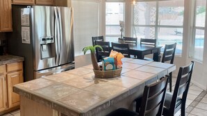 A kitchen with a central island, wooden cabinets, a stainless steel refrigerator, and a dining area with a table and chairs. - ☀️🌵Tropical Desert Oasis. HEATED Pool & Spa no charge. Private, large yard 🌴🏜 (Glendale)