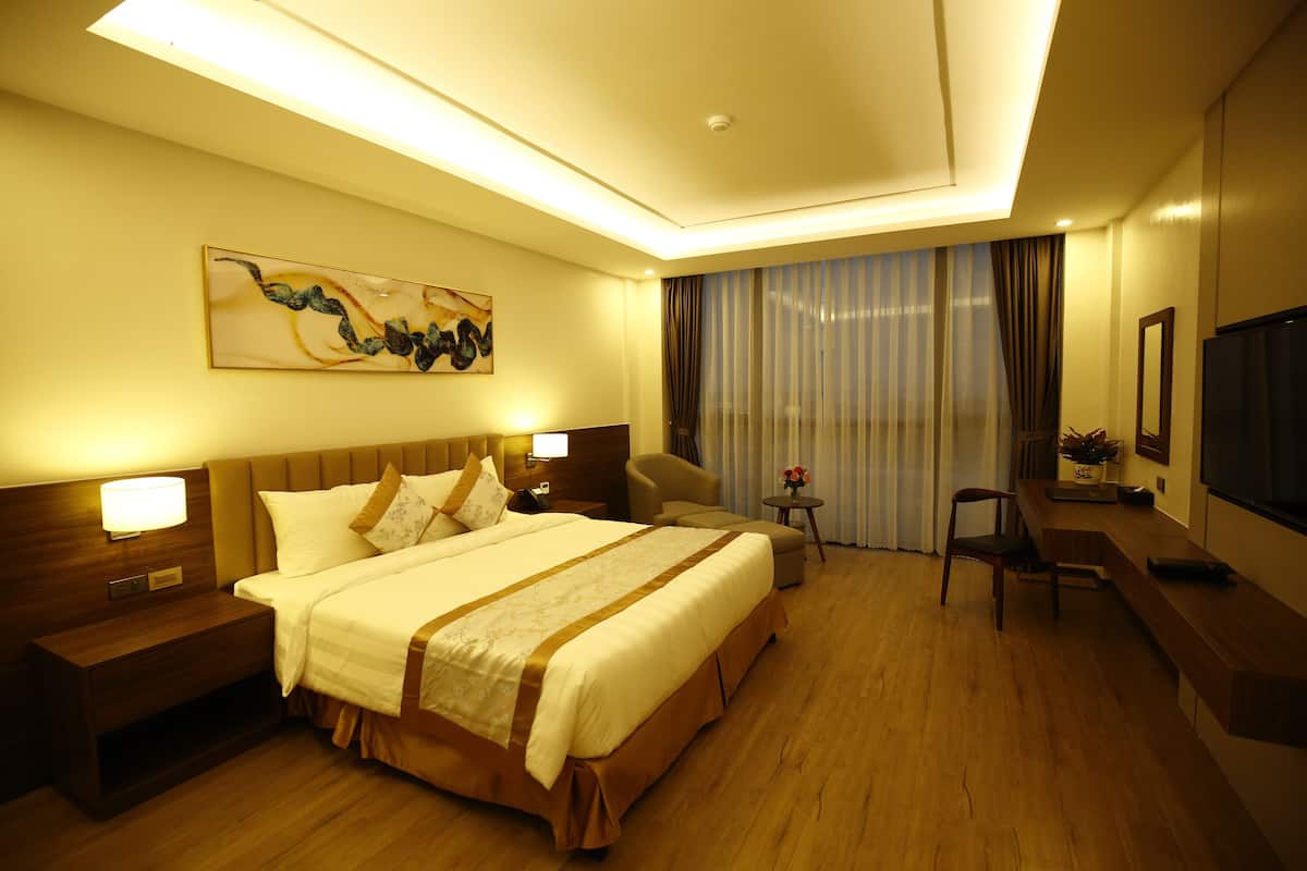 A hotel room with a large bed, bedside tables, a desk, a chair, a TV, and a window with curtains.