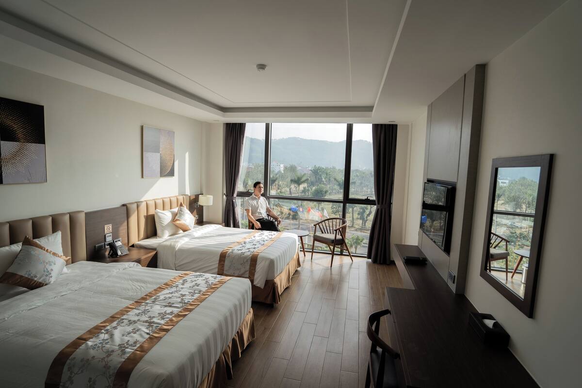 A hotel room with two beds, a desk, and a large window offering a view of the city.