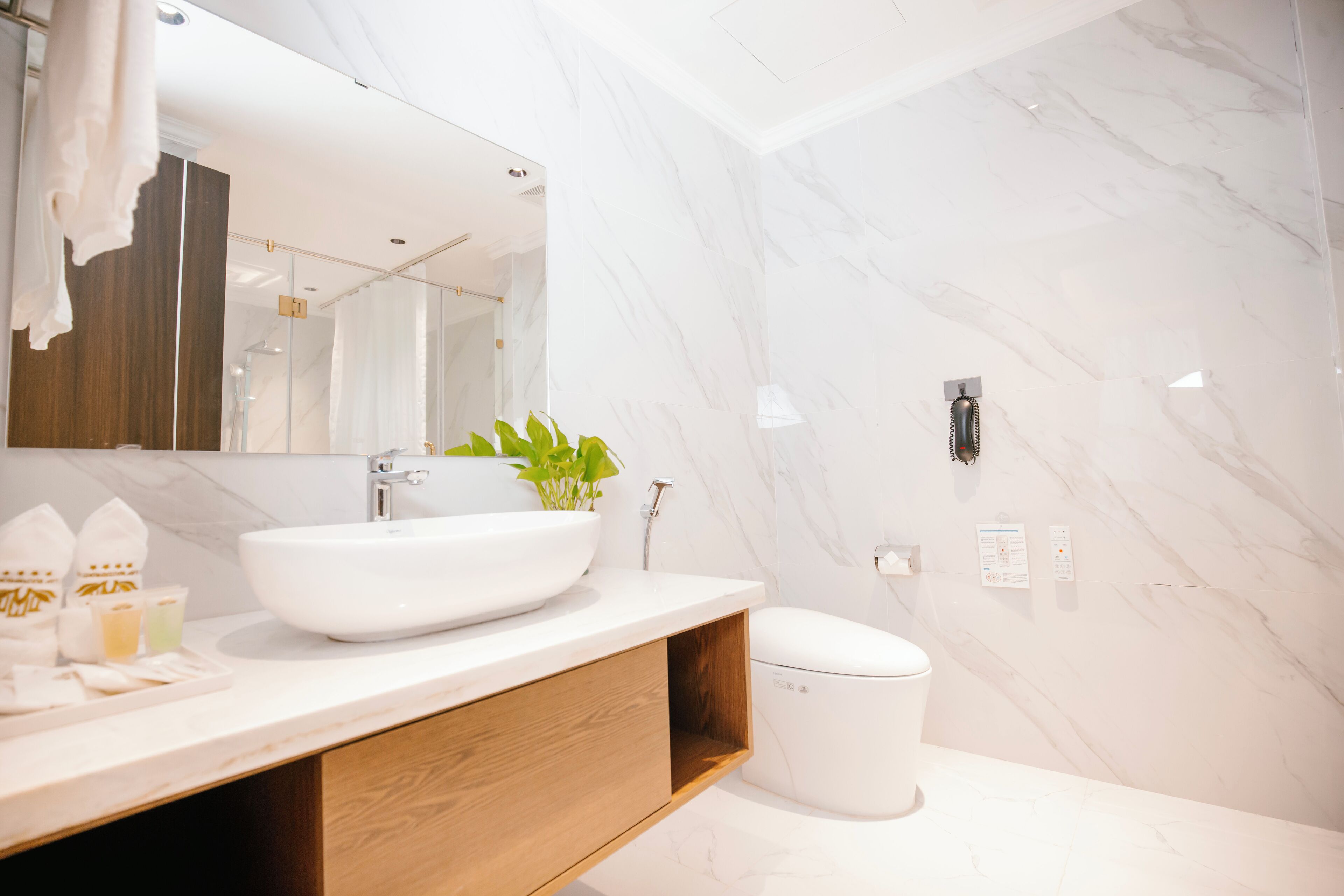 Un baño moderno con un espejo grande, paredes de mármol blanco, un mueble de tocador de madera, un lavabo blanco y un inodoro.