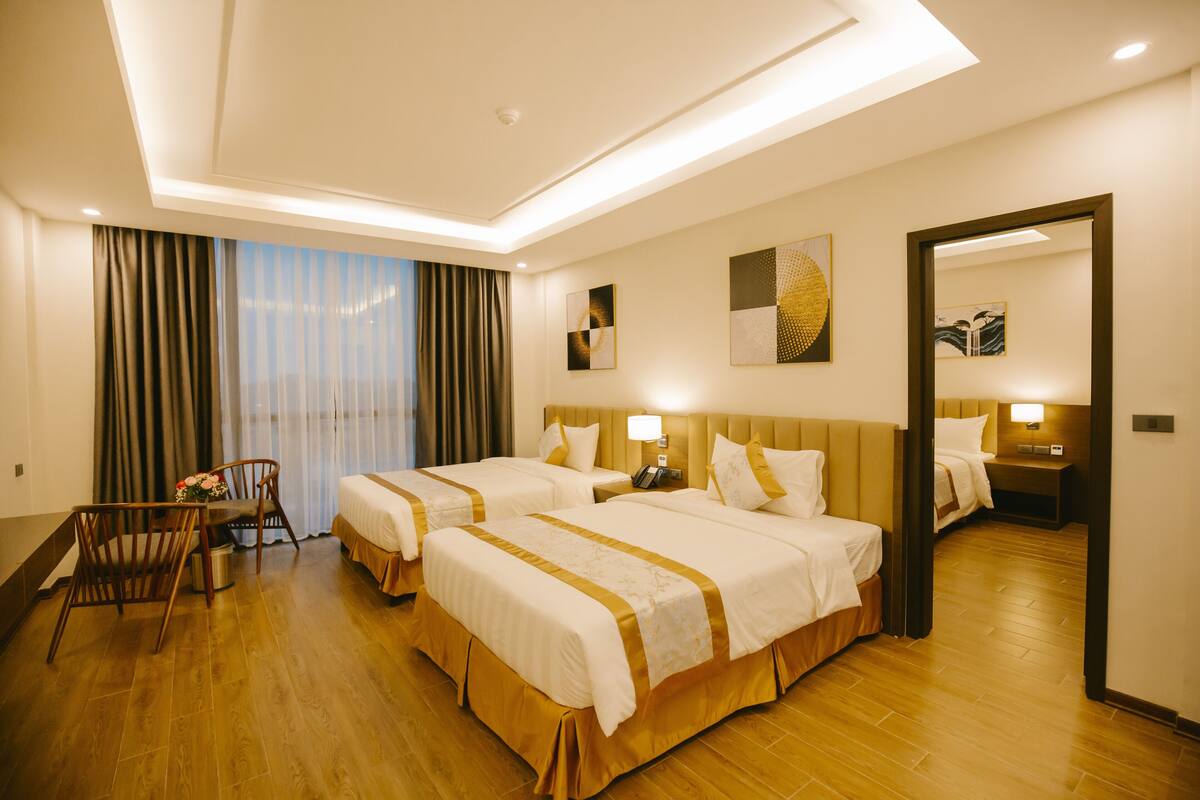 A hotel room with two beds, a desk with a chair, a large window with curtains, and artwork on the walls.