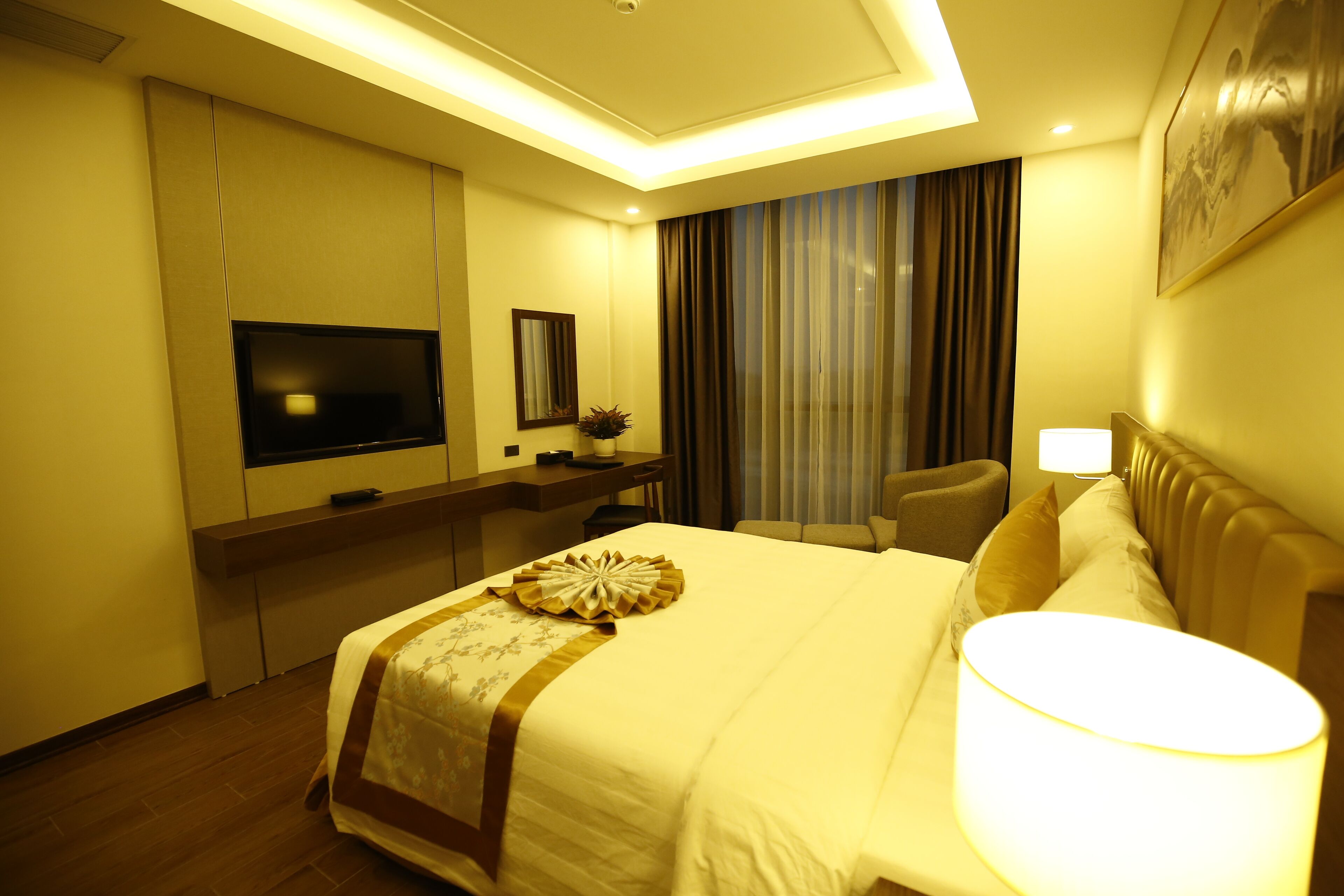 Een hotelkamer met een bed, bedlampje, televisie, bureau en een groot raam met gordijnen.