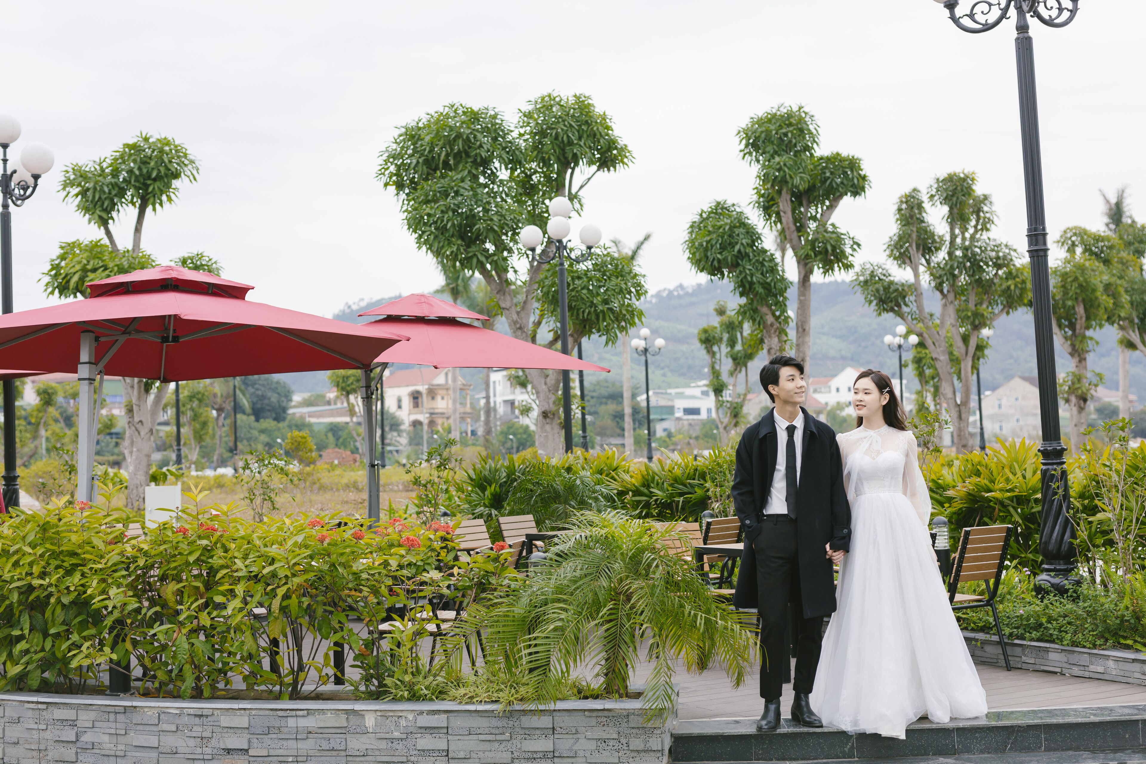 A couple standing in an outdoor area with benches and red umbrellas, surrounded by greenery and trees.