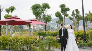 A couple standing in an outdoor area with benches and red umbrellas, surrounded by greenery and trees.
