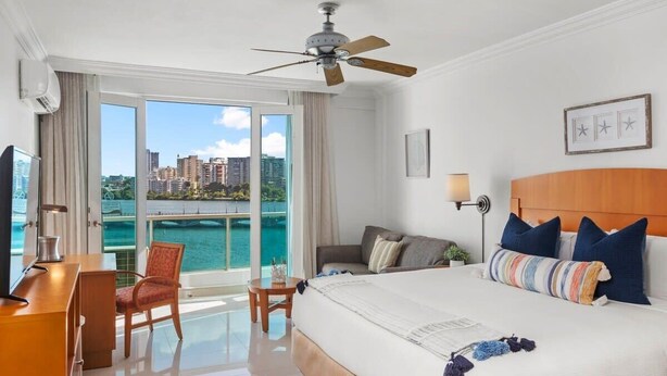Une chambre d’hôtel avec un grand lit, un bureau, une chaise, un ventilateur de plafond et une vue sur une étendue d’eau et des bâtiments.