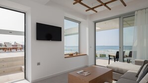 En moderne stue med et fladskærms-TV, et træsofabord og en sofa. Store skydedøre fører ud til en balkon med havudsigt.