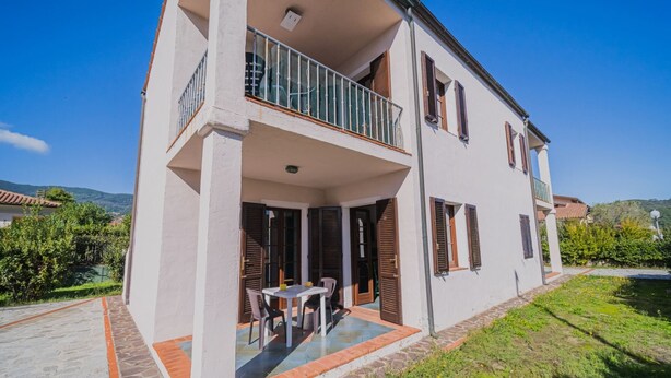 Una casa de dos pisos con un balcón, un patio con mesa y sillas, y un jardín.