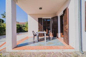Eine Außenpatio mit Tisch und Stühlen, ein Fliesenboden und ein Blick auf einen Garten sowie Gebäude im Hintergrund.