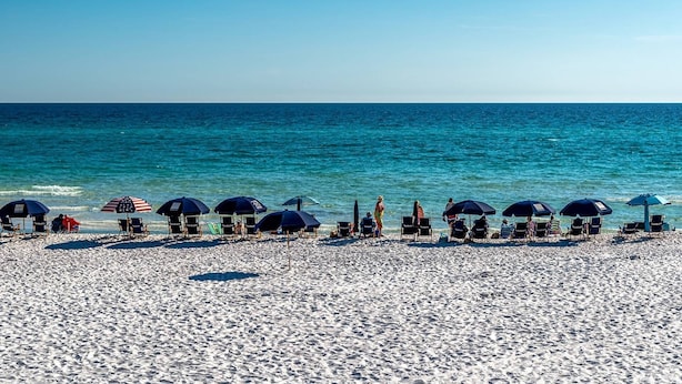 Ein Strand mit Reihen von Liegestühlen und Sonnenschirmen, einem klaren blauen Himmel und ruhiger See.