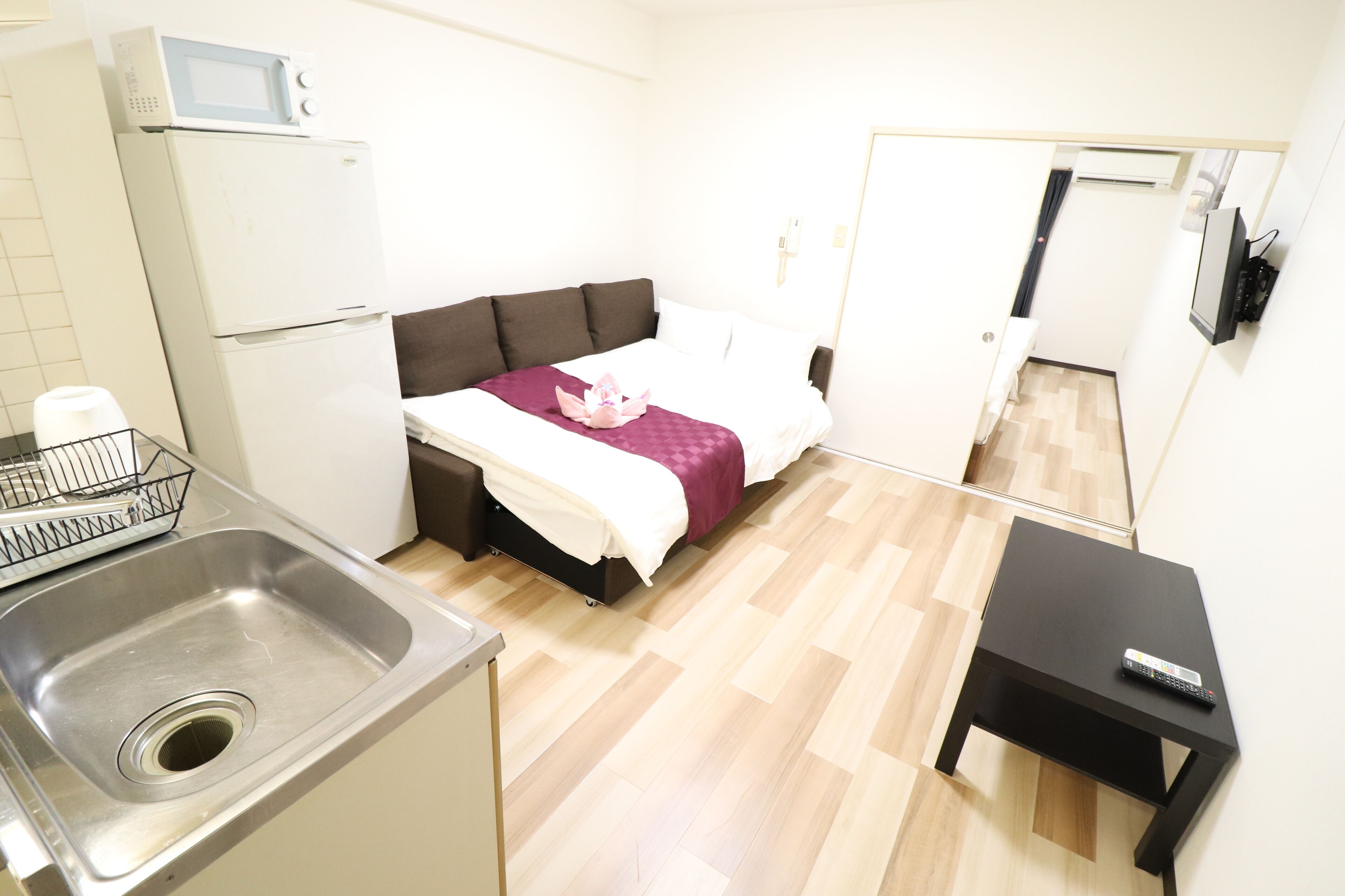 Habitación de hotel compacta con cama, kitchenette pequeña, lavamanos y televisor sobre una mesita.