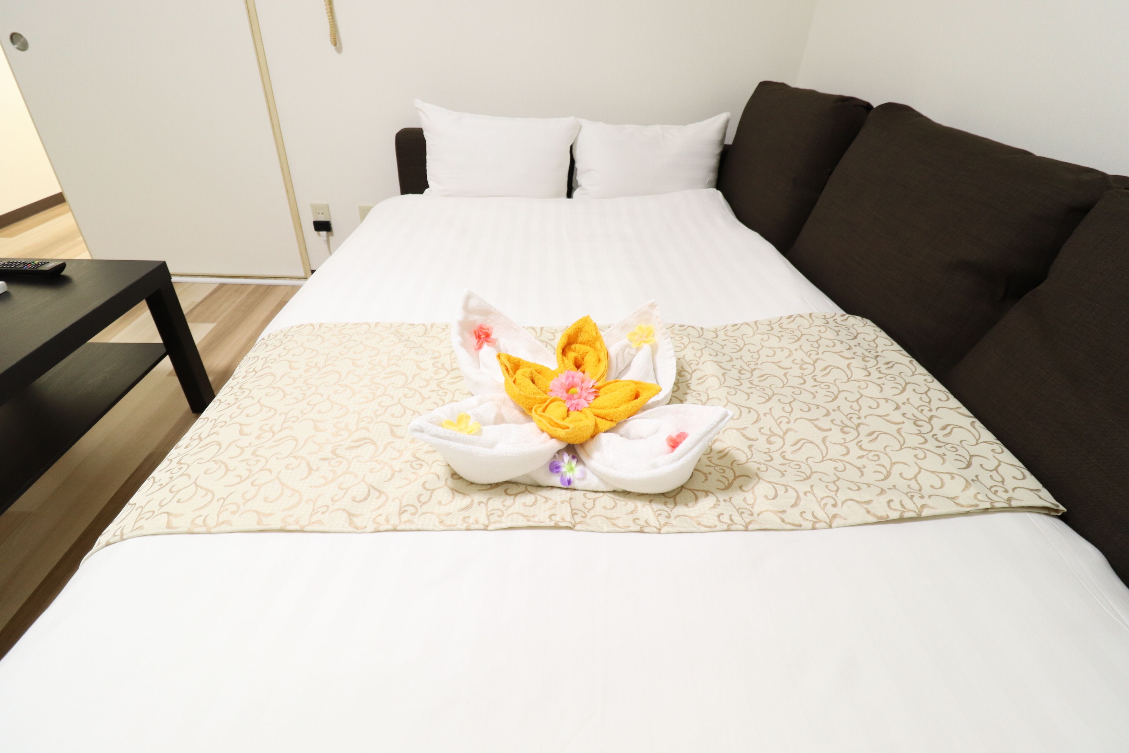 Une chambre d’hôtel avec un lit bien fait, une télécommande posée sur une table de chevet et un canapé marron.