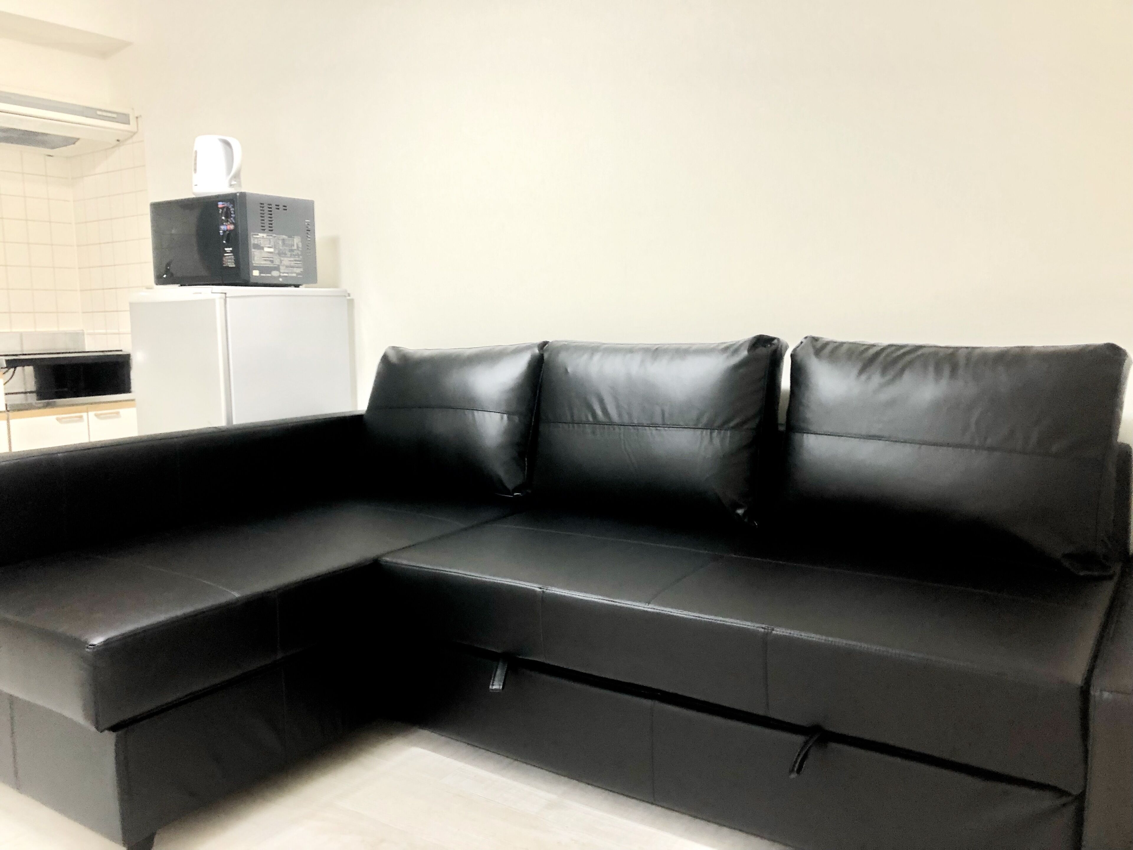 A black leather sofa with a chaise section in a modern kitchen with white cabinets and a microwave.