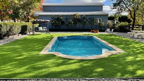 A rectangular swimming pool with a stone border, surrounded by a well-maintained lawn, a patio area with outdoor furniture, and a house with a grey exterior and a sloped roof. - Cosy FarmStay@Harmony Cottage on Melbournes' Famous Mornington Peninsula (Tyabb)