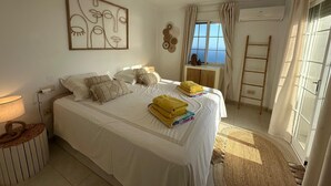 Quarto com cama, ventoinha no teto, janela com cortinas, ar condicionado fixado na parede e mesa de cabeceira.