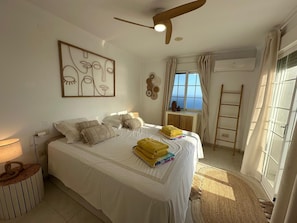 A bedroom with a bed, a ceiling fan, a window with curtains, a wall-mounted air conditioner, and a bedside table.