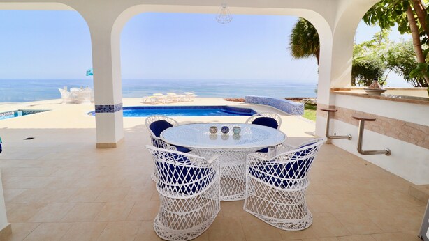 Um pátio com piscina, móveis de vime branco e vista para o mar.