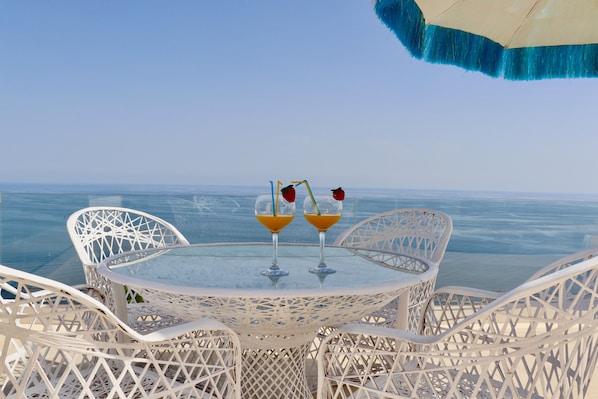 A round table with two glasses of orange drink, surrounded by white wicker chairs, overlooking a body of water.