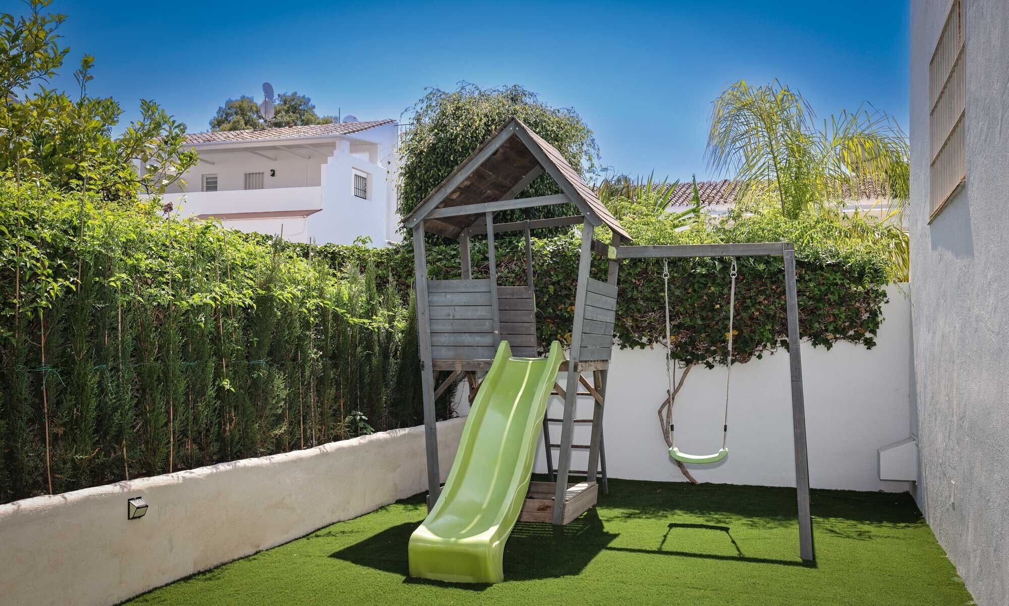 Un giardino sul retro con uno scivolo verde, un'altalena in legno e un muro bianco.