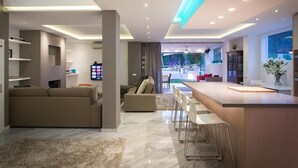 A modern kitchen with a central island, a dining area with bar stools, a living room with a sofa, and a view into the backyard.