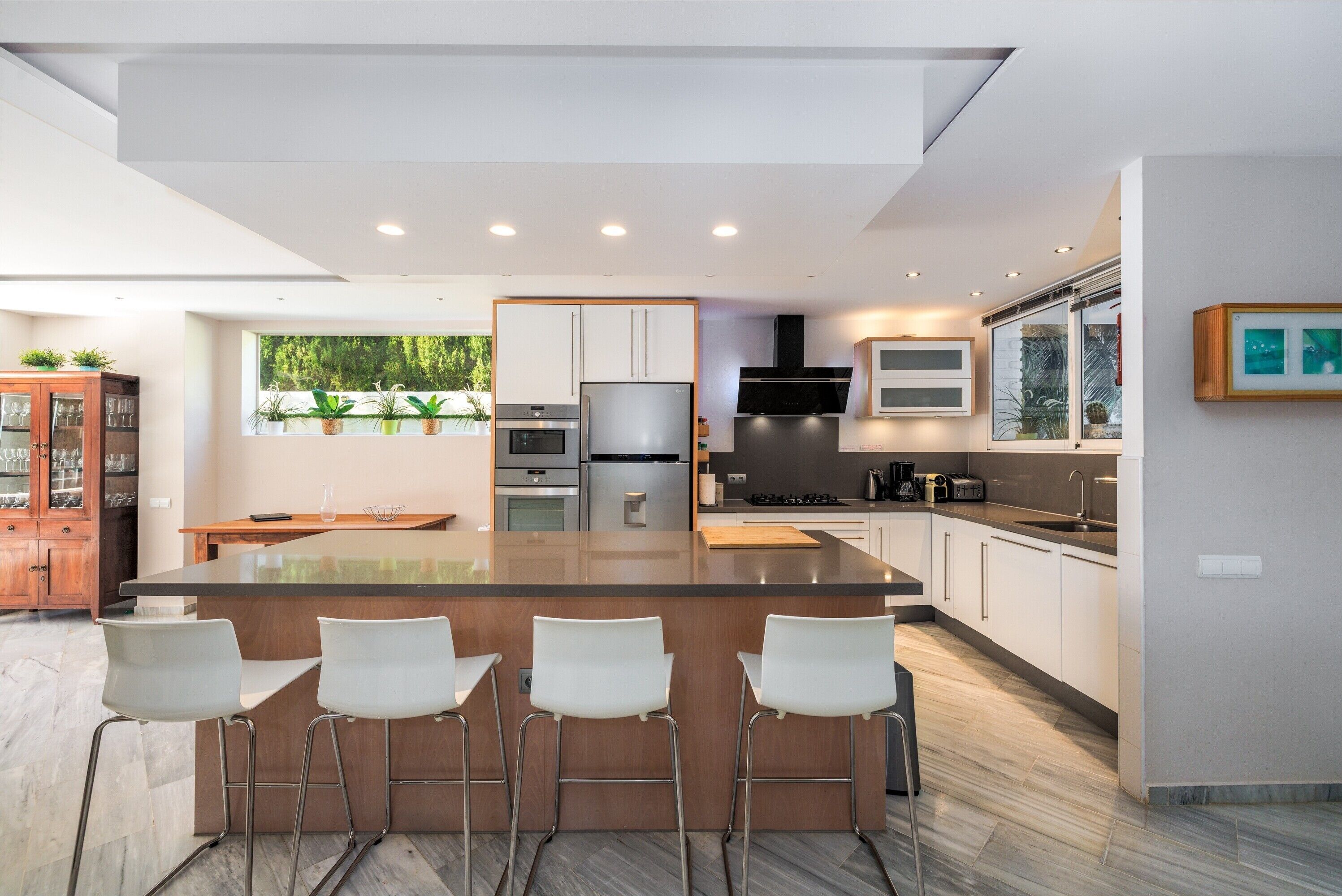 A modern kitchen with a central island, bar stools, stainless steel appliances, and a view of greenery outside.